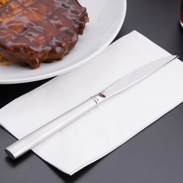 A Libbey Cimarron steak knife on a napkin next to a plate of meat.
