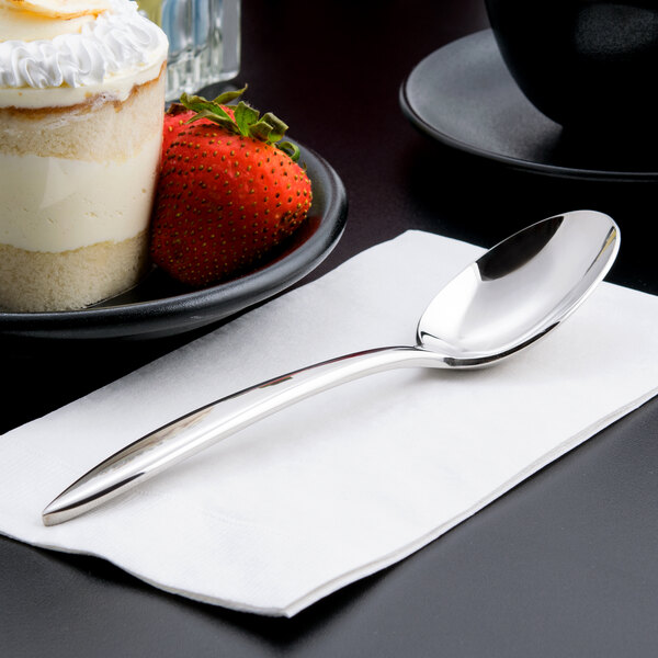 A Libbey stainless steel dessert spoon on a napkin next to a plate of dessert.