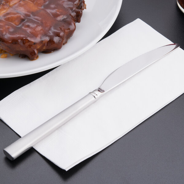 A Libbey Cimarron stainless steel dinner knife on a napkin next to a plate of food.