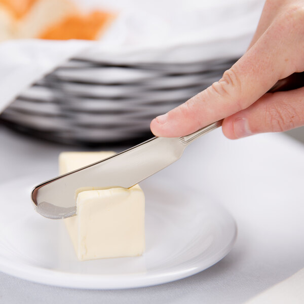 A person's hand holding a Libbey Geneva stainless steel butter spreader over a piece of butter on a plate.