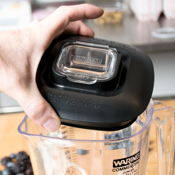 A hand holding a Waring commercial blender with a black plastic lid over a clear container.