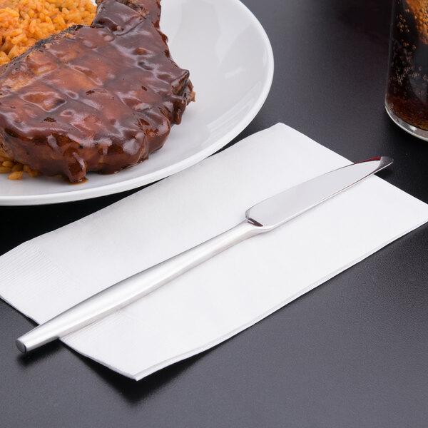 A Libbey stainless steel dinner knife on a napkin next to a plate of meat and rice with a glass of soda.