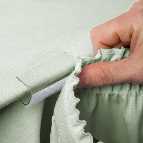 A hand using a Marko plastic hook and loop tape skirting clip to attach white fabric to a table.
