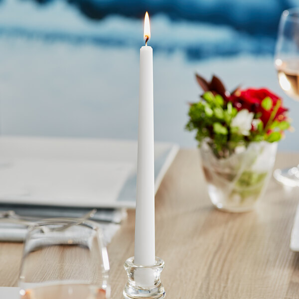 A Will & Baumer white taper candle in a glass holder on a table.