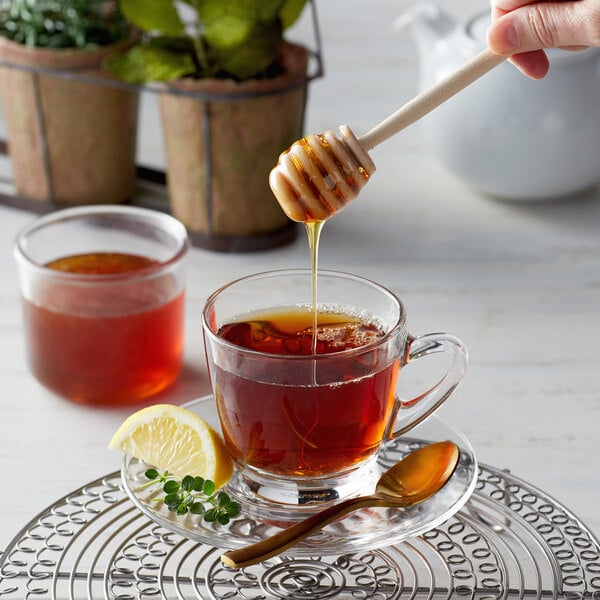 A person using a Monarch's Choice honey dipper to pour honey into a cup of tea.