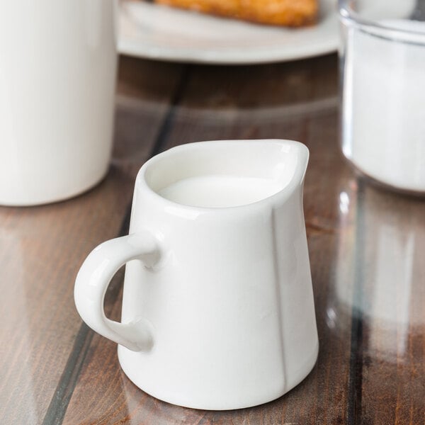 A white Libbey Farmhouse porcelain creamer on a table with a white cup of milk.