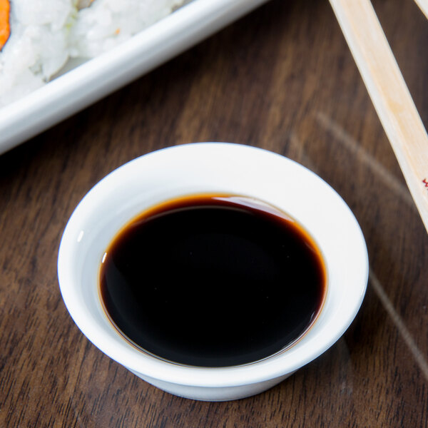 A white Libbey porcelain ramekin filled with soy sauce on a table with sushi and chopsticks.