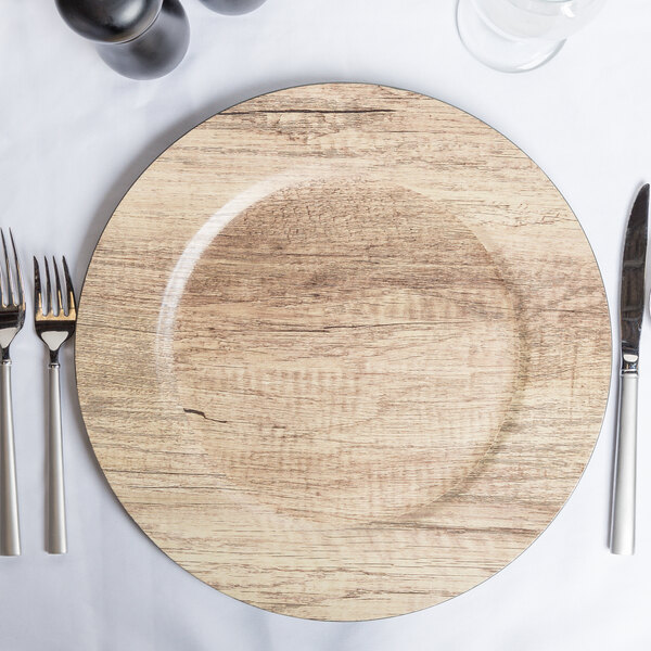 A round faux wood melamine charger plate with silverware and a knife on a white tablecloth.