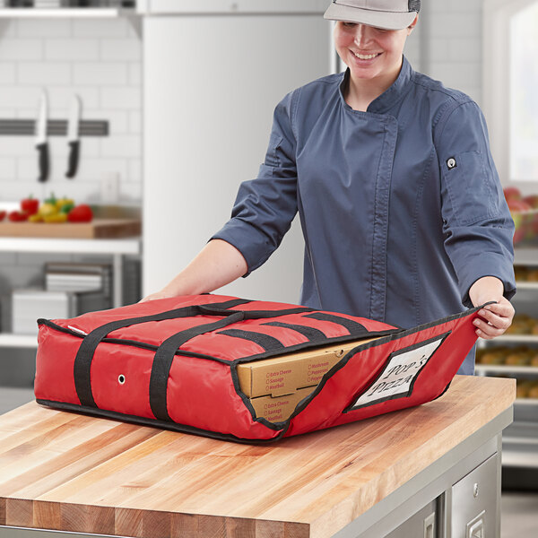 A woman in a chef hat holding a red ServIt insulated pizza delivery bag with a pizza box inside.
