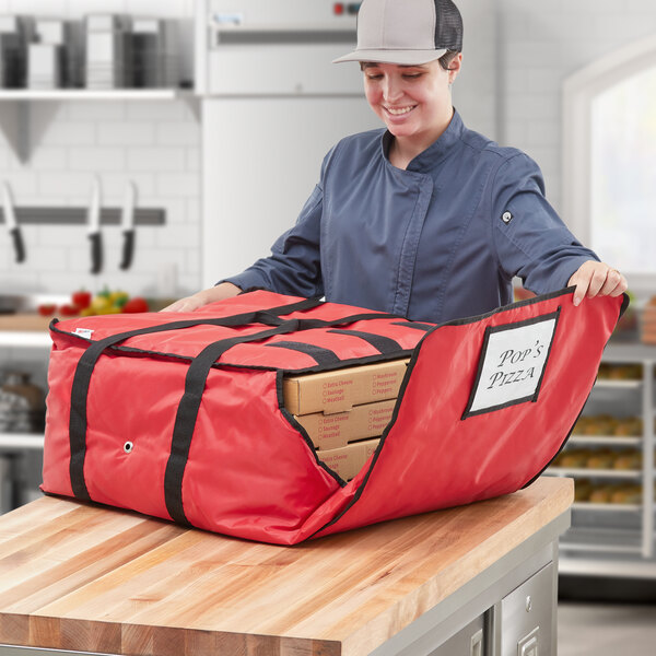 A woman in a chef's hat and blue jacket holding a red ServIt insulated pizza delivery bag with pizza boxes inside.