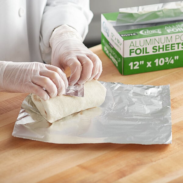 A person in gloves is using Choice interfolded foil sheets to wrap a piece of food on a table.