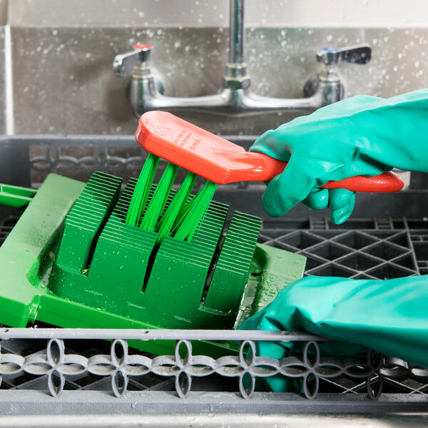 A person in green gloves uses a Prince Castle Saber King cleaning brush to clean a dishwasher.