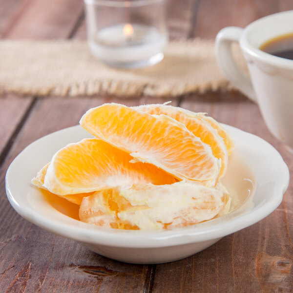 A Tuxton eggshell scalloped edge china fruit bowl filled with orange slices on a table with a cup of coffee.