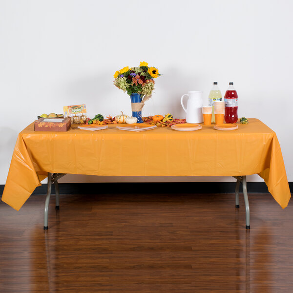 A table with a Creative Converting Pumpkin Spice Orange plastic table cover and autumn flowers on it.
