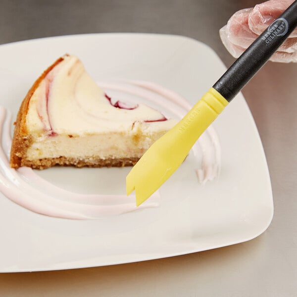 A hand holding a yellow Mercer Culinary saw tooth silicone brush over a slice of cheesecake.