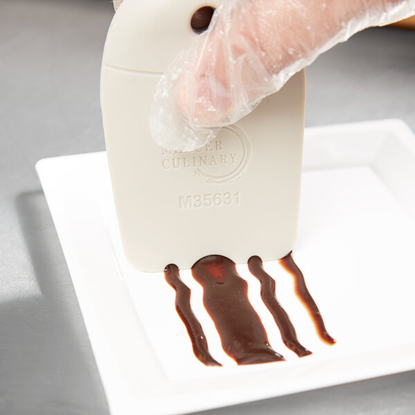 A hand using a Mercer Culinary round arch plating tool to drizzle chocolate syrup over a white rectangular plate of food.