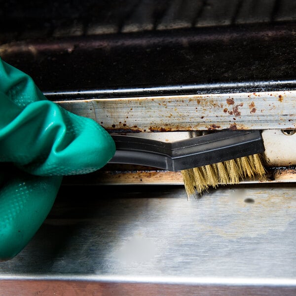 A hand in a green glove using a Winco toothbrush style utility brush with brass bristles to clean a dirty oven.