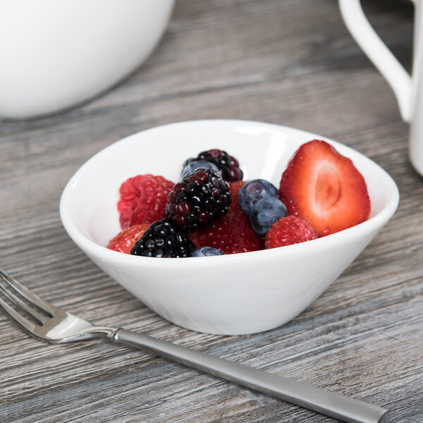 A Libbey white porcelain bowl filled with fruit with a spoon on a wood table.