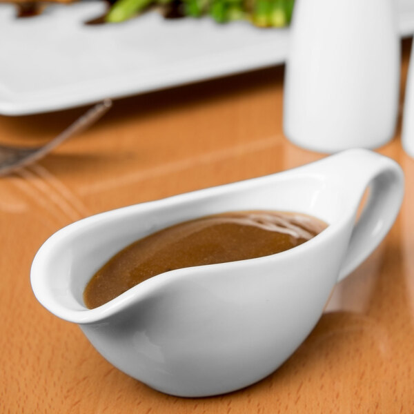 A white Libbey Slenda sauce boat filled with brown sauce on a table.