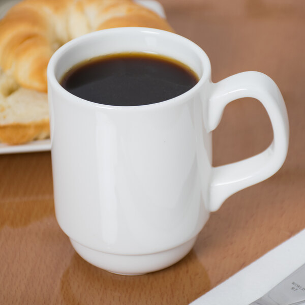 A Libbey white porcelain coffee mug on a table with a bagel and a cup of coffee.