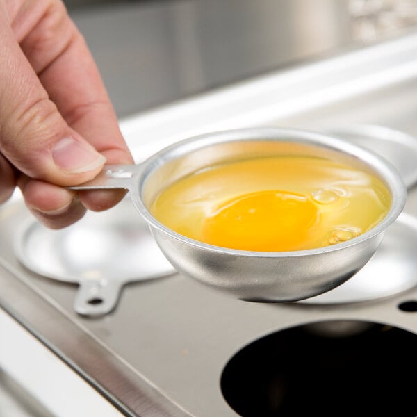 A hand holding a Vollrath aluminum egg poacher cup over a pan with eggs.