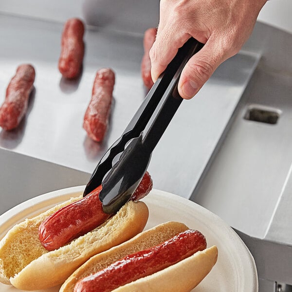 A hand using black Visions tongs to serve a hot dog on a plate.