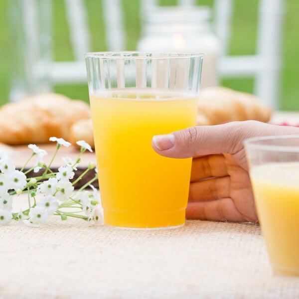 A person's hand holding a WNA Comet tall clear plastic fluted tumbler filled with orange juice.