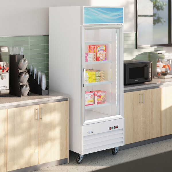 A white glass door merchandiser freezer with LED lighting, stocked with frozen food items and positioned in a break room setting.