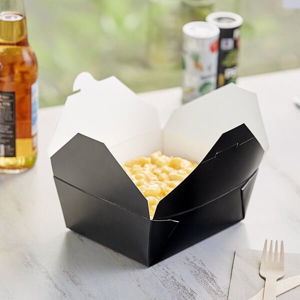 A black Choice microwavable paper take-out box filled with food on a table with a wooden fork on a napkin.