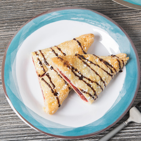 A Kanello melamine plate with two pastries drizzled with chocolate and strawberry syrup on a wood table.
