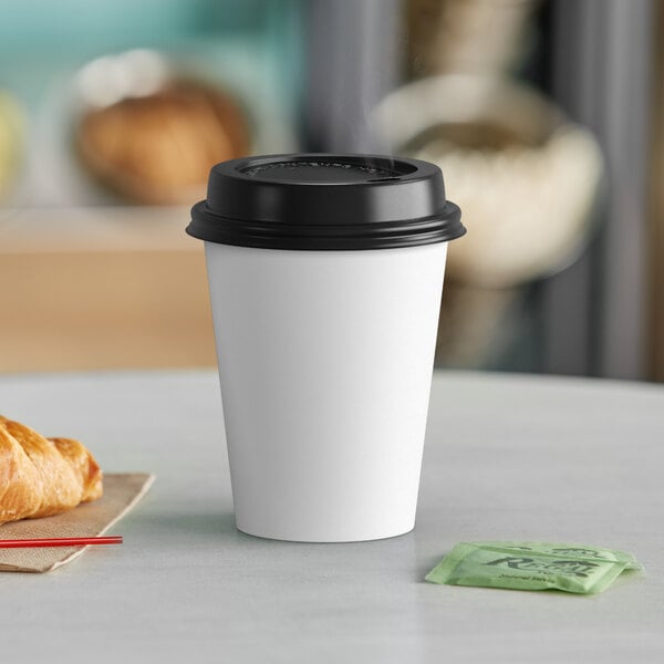 A white Choice paper hot cup with a black lid and a croissant on a table.