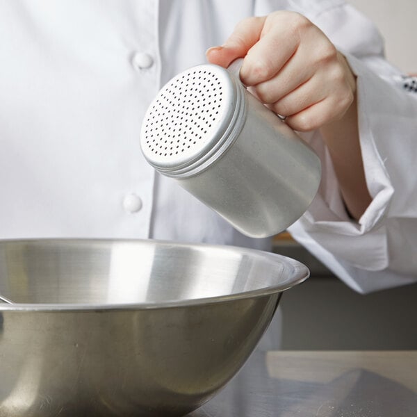 A person in a white chef coat using a Vollrath aluminum shaker with handle to pour something into a bowl.