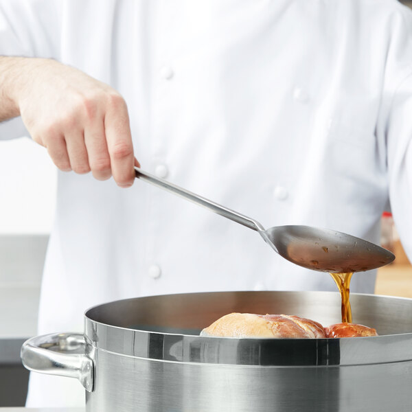 A chef using a Vollrath stainless steel basting spoon to pour brown liquid into a pot of food.