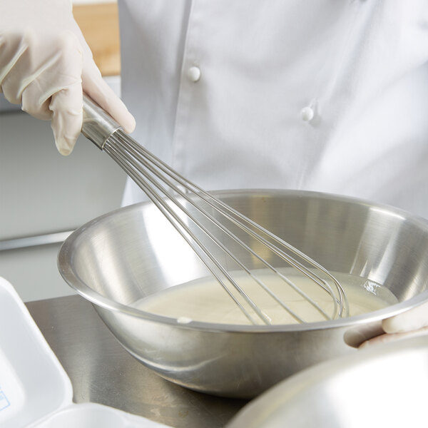 A person in a white chef's coat using a Vollrath stainless steel French whisk to mix a white liquid in a bowl.