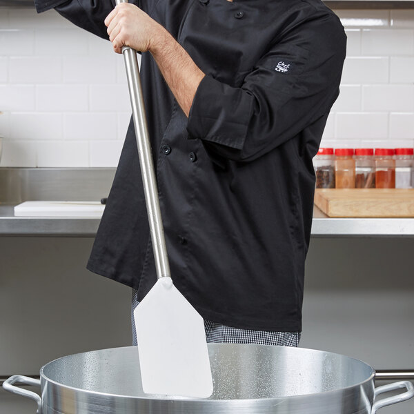 A man in a chef's uniform stirring a large pot with a Carlisle Sparta paddle with a white nylon blade and stainless steel handle.