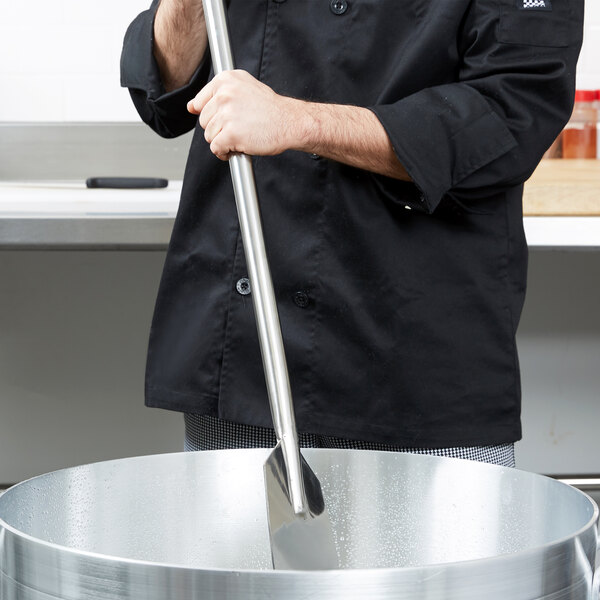 A chef stirring a large pot with a Carlisle stainless steel paddle.