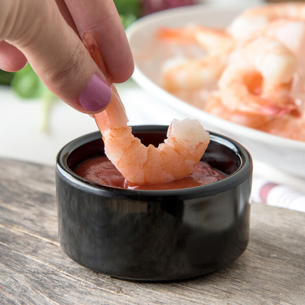 A hand holding a shrimp in a Carlisle ramekin of sauce.