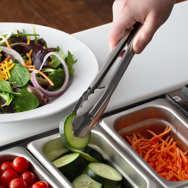 A person using Vollrath stainless steel utility tongs to pick up a cucumber slice from a salad plate.