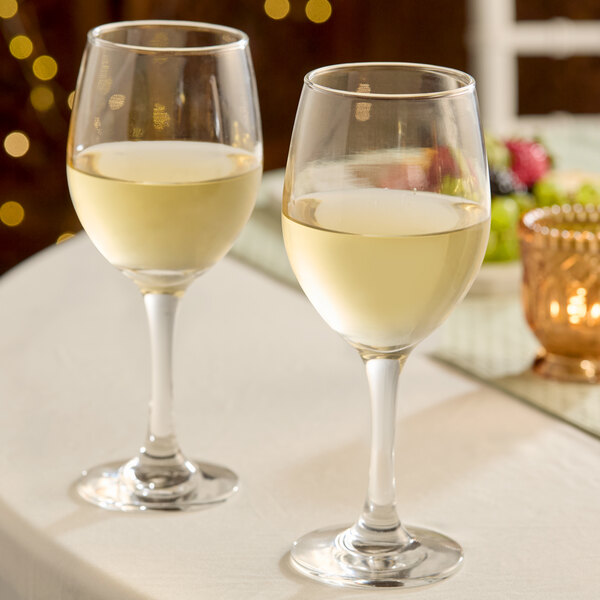 A pair of clear 14 oz. all-purpose wine glasses filled with white wine on a table.