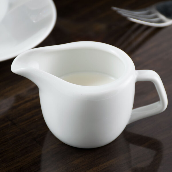A Tuxton bright white china creamer on a table with a white cup and a spoon.