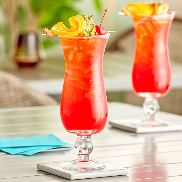 Two Acopa Hurricane glasses filled with red drinks and pineapple garnishes on a table.