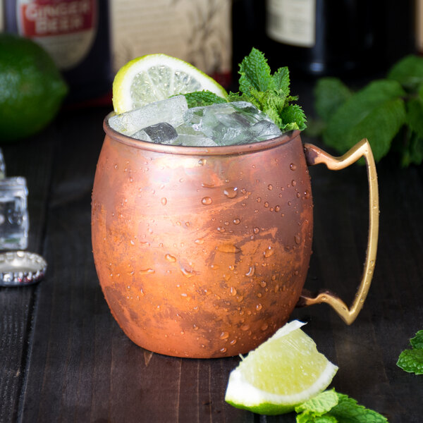 An American Metalcraft copper Moscow Mule mug filled with ice and mint on a wooden surface.