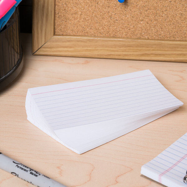 A desk with a stack of Universal white ruled index cards, a notebook, and pens.