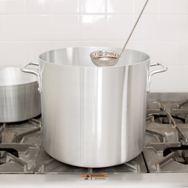 A silver Vollrath Arkadia stock pot on a stove with a ladle on top.