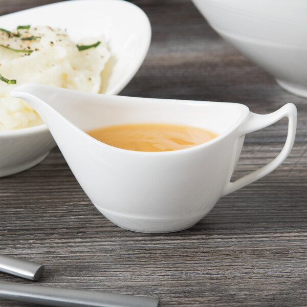 A white Libbey Royal Rideau porcelain sauce boat filled with sauce next to a bowl of mashed potatoes.