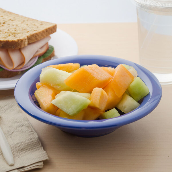 A Carlisle Ocean Blue rimmed melamine bowl filled with fruit and a sandwich on a table.