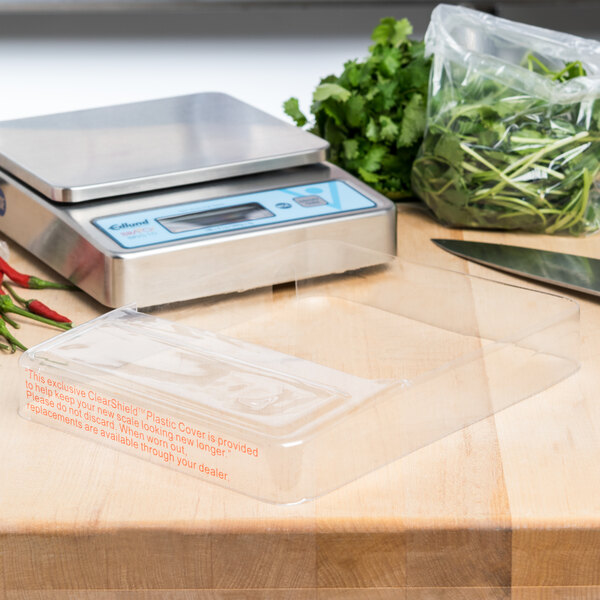 A scale with a plastic container of greens on it with an Edlund ClearShield protective cover.