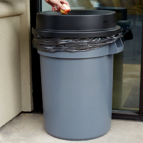 A hand placing an apple into a gray and black round trash can with a black funnel top lid.
