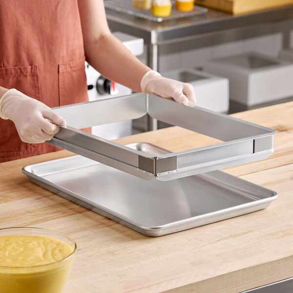 A person wearing gloves is holding a rectangular metal frame above a metal baking sheet on a wooden countertop.