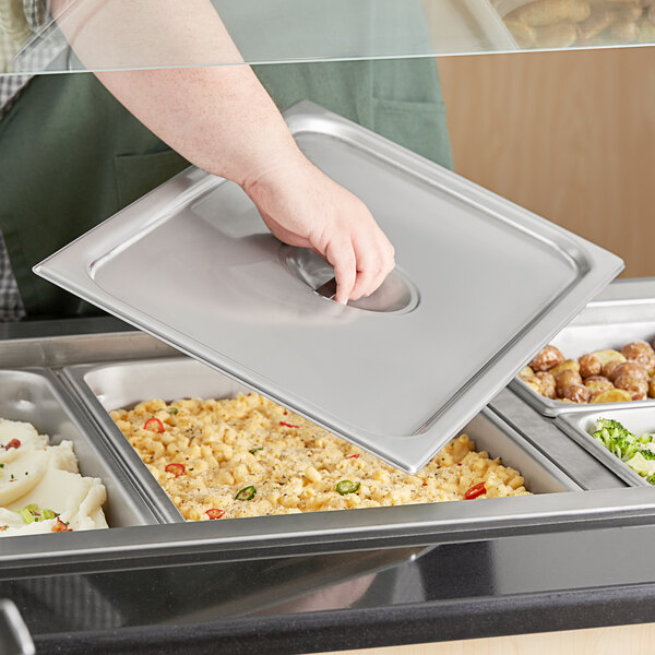 A hand holding a silver Vigor steam table pan cover over a tray of food.